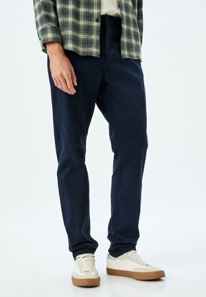 Pantalones de algodón azul marino con un corte de pierna recta, combinados con una camisa de franela de cuadros verdes y zapatillas blancas, que presentan una superficie texturizada.