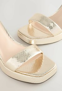 Arany metál magas sarkú szandálok négyzet alakú nyitott orral és széles elülső pántokkal, melyeken texturált, kígyóbőr-hatású minta látható.