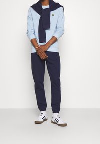 Sudadera de color azul claro con detalles en navy, combinada con pantalones de chándal navy. Zapatillas blancas con franjas en navy y negro. Presenta un pequeño logo en las prendas.