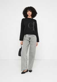 Melnas, īsas džemperis ar pelēku logo, kopā ar pelēkām, ar augstu jostasvietu un platajām biksēm. Modele tur nelielu melnu somu un valkā melnas dzīvokļu kurpes.