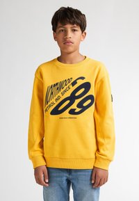 Sudadera amarilla con cuello redondo, presenta un gran texto gráfico negro y un logo. La tela tiene una textura suave. Ajuste casual.