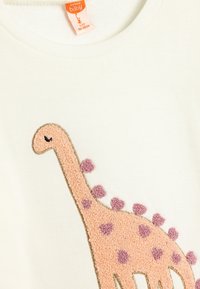 Witte babytrui met lange mouwen, featuring een gestructureerd, roze dinosauruss ontwerp met paarse vlekken en een gouden rand. Het etiket toont maat 104-110cm.