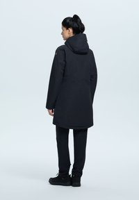 Svart vattentät parka med huva, långa ärmar, sidofickor och rak passform, kombinerad med svarta byxor och skor. Slät yta.