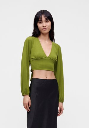 Kvinde med sort hår iført en grøn langærmet crop top og en sort højtaljet nederdel, står foran en ensfarvet lyse baggrund.