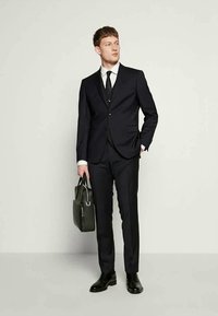 Noormees mustus ülikonnas, valges särgis ja mustas lipsus, hoides musta nahast portfelli, seistes ühevärvilise heledat tausta vastu.