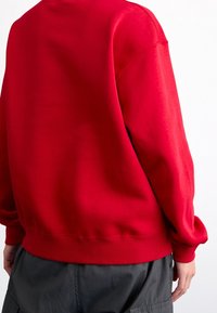 Person trägt ein hellrotes Sweatshirt und dunkelgraue Cargo-Hose, von hinten vor einem schlichten weißen Hintergrund gezeigt.