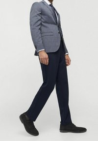Uomo che cammina indossando un blazer a quadri blu, una camicia bianca, una cravatta nera, pantaloni blu scuro e scarpe in suede nere su uno sfondo chiaro e semplice.