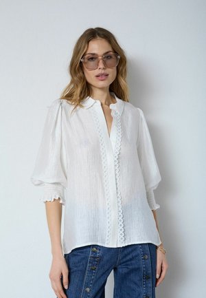 Vrouw met oversized getinte bril, een witte blouse met structuur en kanten afwerking en opgeblazen mouwen, gecombineerd met een blauwe broek met knoopdetails.