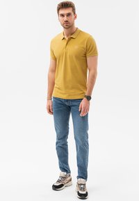 Polo giallo con maniche corte e un logo sul petto, abbinato a jeans di denim blu e sneakers multicolori.
