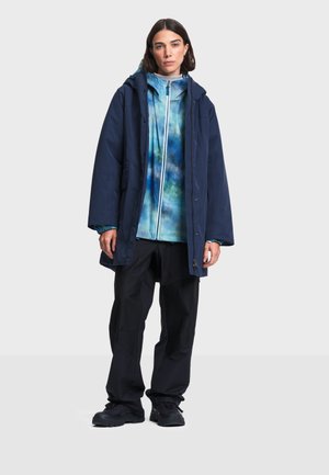 Kvinde iført en marineblå hættejakke over en blå og hvid tie-dye lynlåstrøje, sorte bukser og sorte sko, stående mod en lys baggrund.