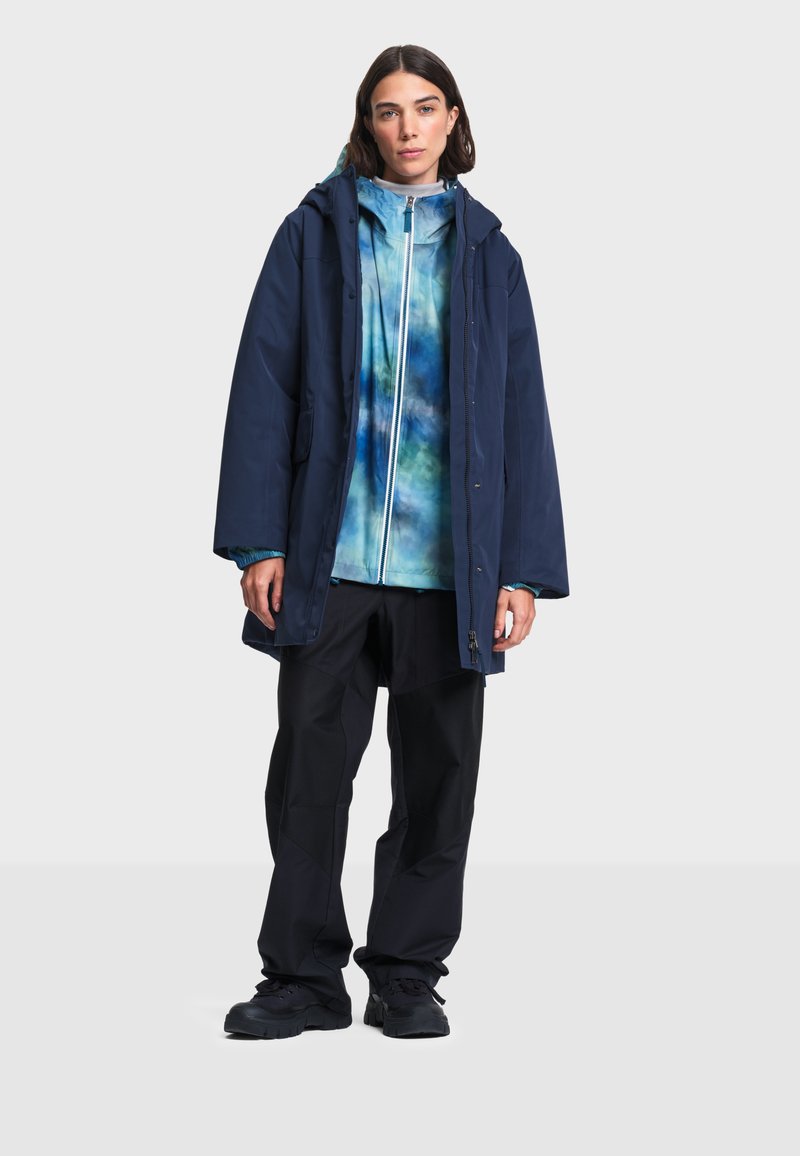 Femme portant une veste à capuche bleu marine par-dessus une veste zippée tie-dye bleue et blanche, pantalon noir et chaussures noires, debout devant un fond clair.
