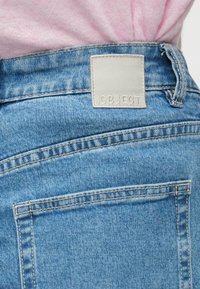 Svetlo modra jeansa tkanina, ki vključuje zadnji žep, pravokotni sivi etiketi z besedilom "OBJECT" in vidnimi podrobnostmi šivanja.