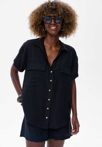 Czarna krótka koszula z guzikami, z dwoma kieszeniami z przodu, z drewnianymi guzikami i luźnym fasonem. Noszona z pasującymi szortami.