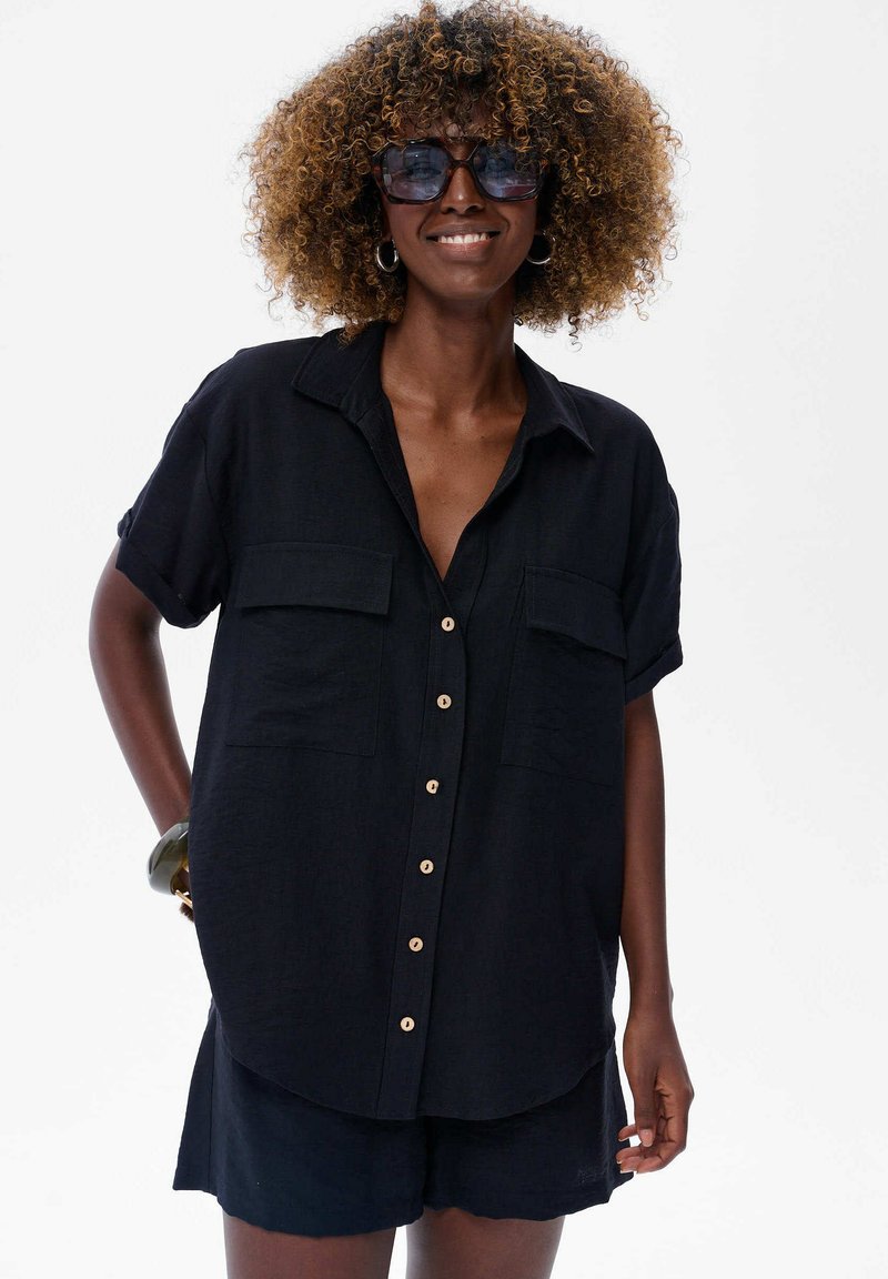 Czarna krótka koszula z guzikami, z dwoma kieszeniami z przodu, z drewnianymi guzikami i luźnym fasonem. Noszona z pasującymi szortami.