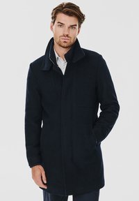 Granatowy wełniany płaszcz z wysokim kołnierzem, zapięciem na zamek i kieszeniami bocznymi; charakteryzuje się smukłym krojem i miękką strukturą.