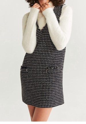 Kobieta w czarno-białej, fakturowanej sukience bez rękawów na długim, puszystym, białym swetrze z rękawami, ręce przy szyi, nogi bez zakrycia.