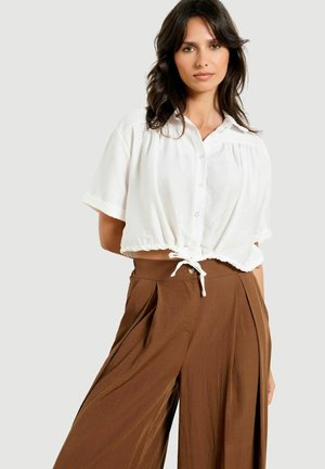Młoda kobieta o ciemnych włosach nosi białą, krótką bluzkę typu crop top z guzikami oraz szerokie, brązowe spodnie z wysokim stanem na jednolitym tle.
