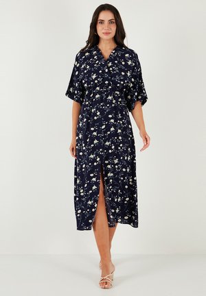 Abito lungo floreale blu navy con maniche corte, chiusura a bottoni e spacco laterale. Presenta un tessuto leggero con motivi di fiori gialli e bianchi.