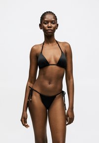 Γυναίκα με πλεξούδες που φοράει μαύρο μπικίνι δύο τεμαχίων με κορδόνια, στέκεται μπροστά σε σκουρόχρωμο γκρι φόντο.