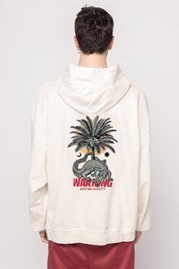 Cremefarbenes, übergroßes Hoodie mit einem grafischen Design eines Palmbaums, eines Auges und eines Krokodils. "WARNUNG" in roter Schrift darunter.