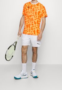 Camiseta atlética de patrón naranja de Puma, pantalones cortos blancos, zapatillas deportivas blancas con acentos en verde azulado, sosteniendo una raqueta de tenis negra y verde.