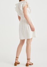 Robe blanche avec des épaules à volants, taille smockée et jupe fluide. Portée avec des sandales à talons hauts striées brunes.