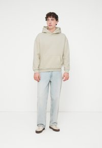 Φόρμα με κουκούλα σε ανοιχτό μπεζ χρώμα με τσέπη καγκουρό, συνδυασμένη με φαρδιά ανοιχτό μπλε τζιν. Η εμφάνιση έχει άνετη εφαρμογή και ελάχιστες σχεδιαστικές λεπτομέρειες.