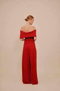 Ženska z blond lasmi, z nizkim čopom, oblečena v rdež kombinezon brez naramnic s širokimi hlačami, stoji ob bež ozadju in se obrača stran.