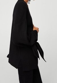 Cardigan noir oversize avec de longues manches bouffantes. Tissu en maille texturée, noué à la taille, fentes sur les côtés pour faciliter les mouvements.