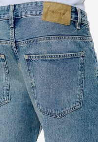 Jeans din denim de culoare albastru deschis, având un design clasic cu cinci buzunare, cusături vizibile și o etichetă din piele maro cu branding-ul "PULL&BEAR" pe talie.