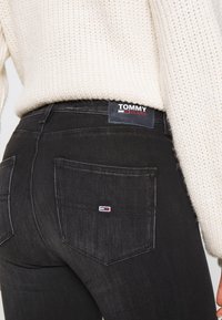 Jeans de denim negro con un diseño ajustado, que presentan una etiqueta de marca en la parte trasera y un pequeño detalle de logo en el bolsillo trasero. Suéter de punto texturizado encima.