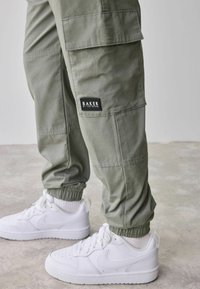 Olijfgroene cargobroeken met elastische boorden, voorzien van zijzakken en een rechthoekig merketiket. Gepaard met witte, lage sneakers.