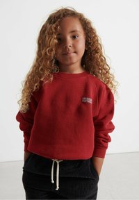 Raudona džemperis su tekstūru ir giliu rankogaliu, puošiamas "AMERICAN VINTAGE" logotipu. Derinamas su tamsiomis aksominėmis kelnėmis.