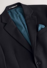Veste de costume noire avec doublure intérieure bleue à motifs et pochette bleue assortie visible dans la poche poitrine.