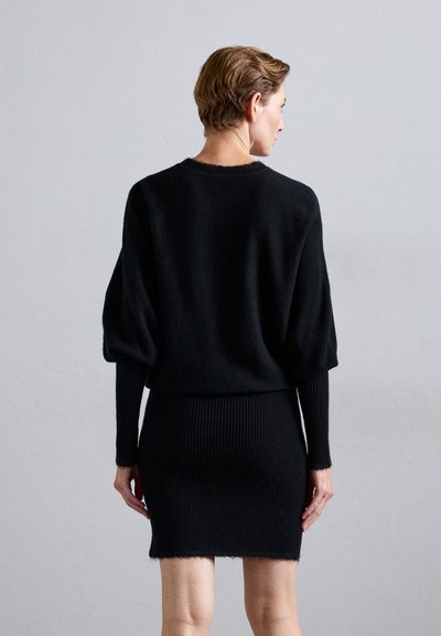 Donna di spalle che indossa un maglione nero a maniche lunghe con maniche a sbuffo e una gonna a tubino nera a coste su uno sfondo grigio neutro.