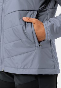 Giacca isolante grigio chiaro con texture trapuntata, chiusura frontale con zip e tasche laterali. Il tessuto è liscio e leggermente lucido.