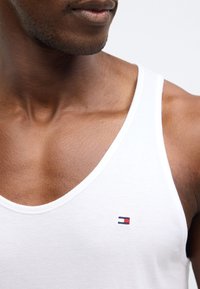 Maiou alb din bumbac cu decolteu adânc în V și un mic logo brodat cu accente roșii, albe și albastre pe partea stângă a pieptului.