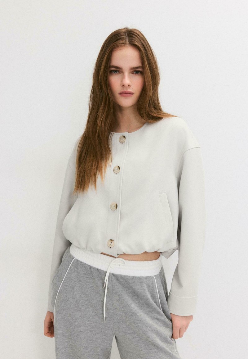 Jeune femme aux longs cheveux bruns portant une veste blanche à boutons et un pantalon de survêtement gris à cordon, sur un fond blanc uni.