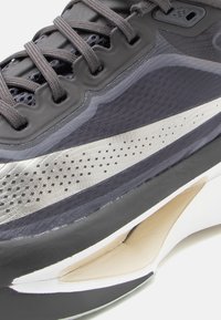 Sportskor med en svart och grå mesh-övre del, med silverfärgad accentlinje, texturerade detaljer och en vadderad vit sula.