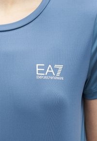 Κοντινό πλάνο ενός ατόμου που φοράει ανοιχτό μπλε μπλουζάκι με το λευκό λογότυπο "EA7 Emporio Armani" στο στήθος.