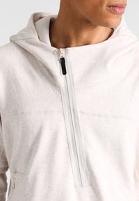 Sudadera ligera gris claro hecha de tela suave, con un cierre central de cremallera y capucha con cordón. Detalles texturizados a lo largo de los hombros.