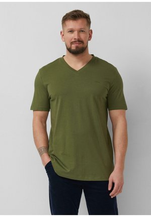 Mężczyzna z brodą, ubrany w oliwkowo-zielony T-shirt z dekoltem w serek i ciemne spodnie, stojący z jedną ręką w kieszeni na jednolitym tle.