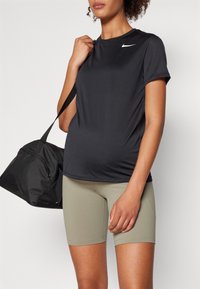 Musta urheilupaita, jossa on lyhyet hihat ja valkoinen logo, yhdistetty kapeisiin oliivinvihreisiin shortseihin. Musta duffel-laukku pidetään yhdessä kädessä.