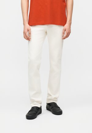 Egy személy vékony szabású törtfehér nadrágot, fekete csúszásmentes cipőt és narancssárga inget visel, egy egyszerű világos háttér előtt áll.