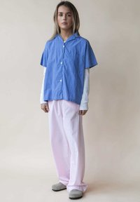 Blauw gestreept overhemd met korte mouwen en een kraag, gecombineerd met lichtroze wijde broek. De outfit wordt aangevuld met grijze instapschoenen.
