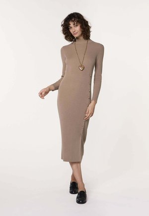 Vrouw die een beige geribbelde midi-jurk met lange mouwen en zijknopen draagt, zwarte instapschoenen en een ketting met hartvormige hanger, ontspannen staand.