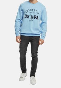 Felpa azzurra con testo stampato nero che dice "AUTHENTIC US PA" e un logo, abbinata a jeans scuri e sneaker bianche.