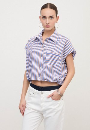Donna che indossa una maglietta corta a maniche corte a righe blu, marroni e bianche con una tasca frontale e pantaloni bianchi con una fascia nera in vita visibile.
