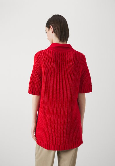 Červený pletený oversized polo top s krátkymi rukávmi, so štruktúrovaným vzorom a rebrovaným golierom. Nosený cez béžové nohavice.