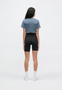 Donna con lunghi capelli scuri vista di spalle, indossa un top corto blu, pantaloncini da ciclismo neri a vita alta, calzini bianchi e scarpe da ginnastica bianche.